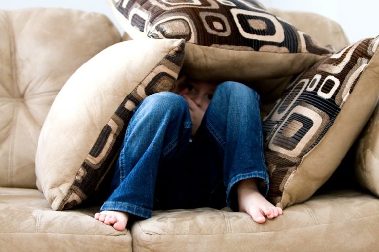Toddler on couch hiding under pillows, color photo