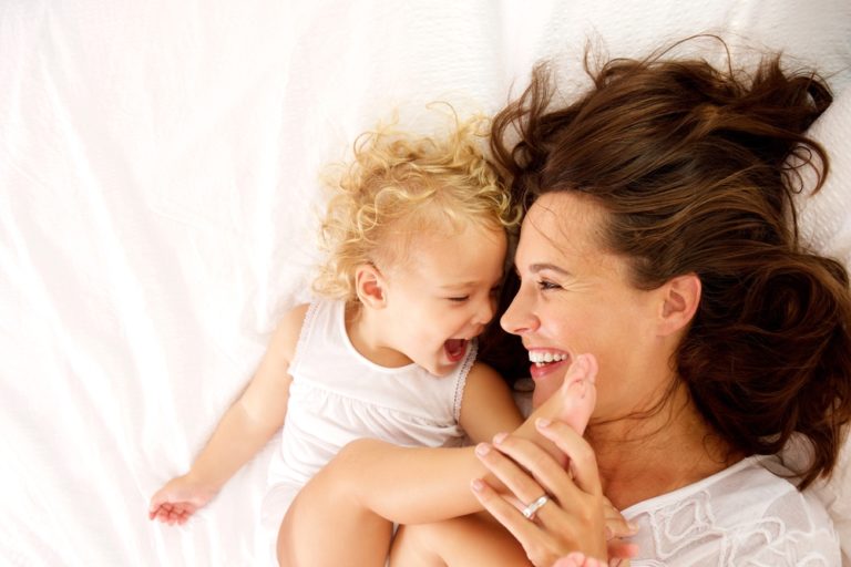 mom and toddler laugh on a bed at home