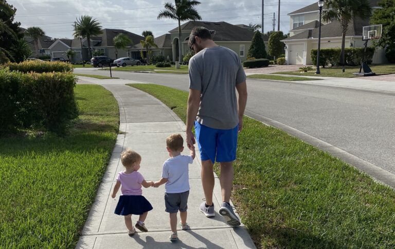 Daddy walking down sidewalk with toddler son and daughter, color photo
