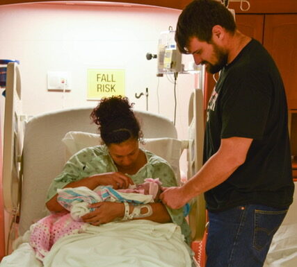 Wife holding stillborn baby with husband standing beside her, color photo