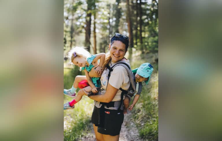 Mother holding a toddler with an infant in a carrier on her back, color photo