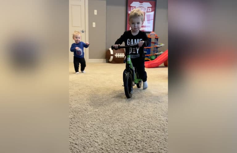 Little boy riding bike in family room with little brother following, color photo