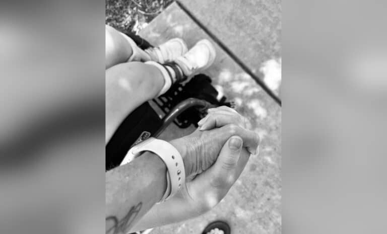 Two hands held together next to a wheelchair, black-and-white photo