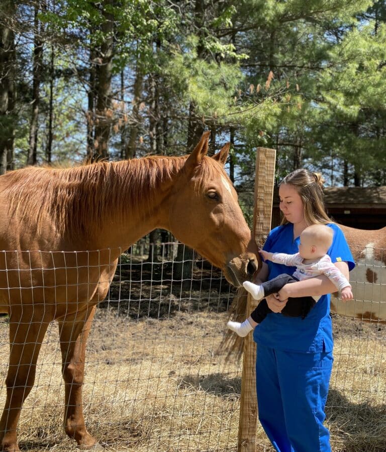 woman in scrubs holding baby and patting horse