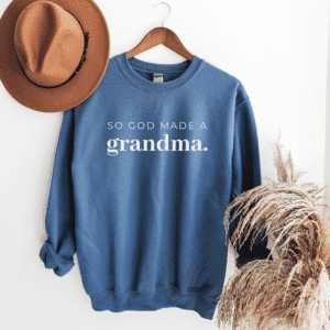 Blue sweatshirt with long sleeves and lettering across the chest reading So God Made a Grandma, in a flat lay with brown hat and tan feathers.