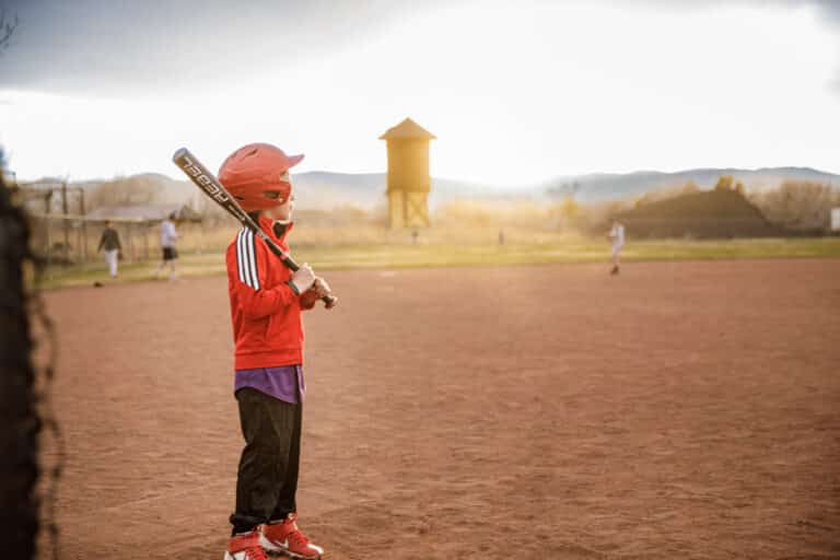 Little boy in a red shirt on sandlot field holding a bat with sun shining in the background