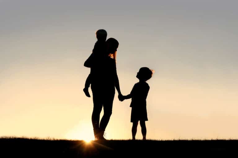 Mother holding hand of young child while holding toddler, silhouette against sunset