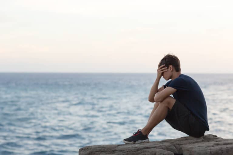 Teen boy sitting on rock over ocean with head in hands