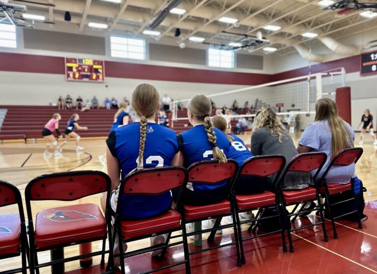 Volleyball team playing in high school gymnasium, girls in blue jerseys on a bench in foreground