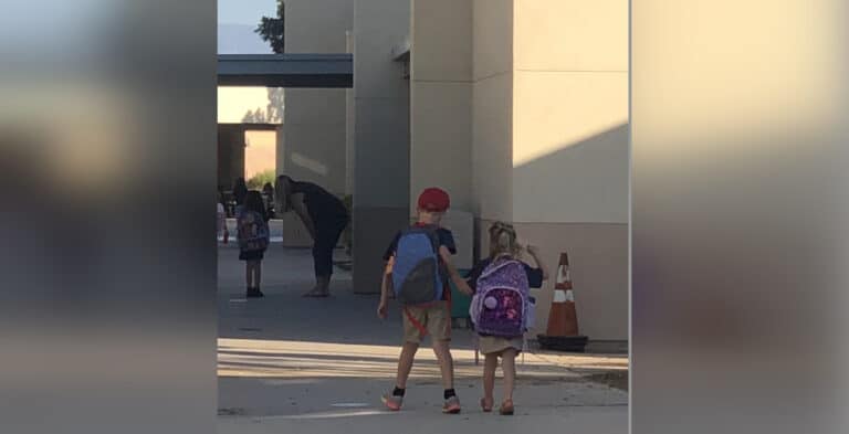 Two young children in backpacks walk toward a school building