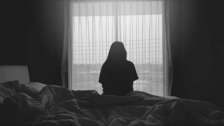 Woman sitting on edge of bed looking out window, black and white image