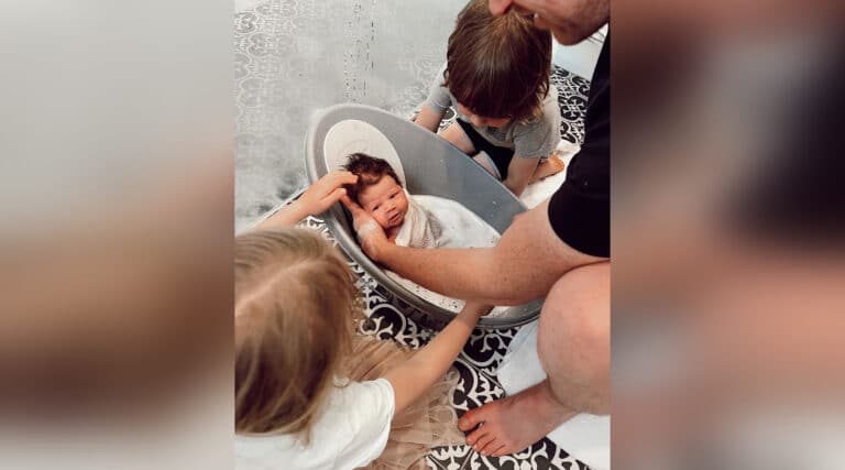 Father holding newborn in bathtub with siblings nearby