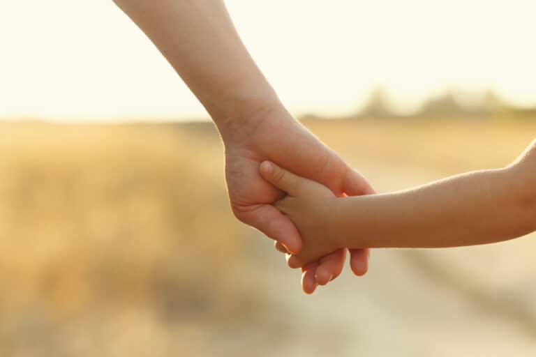 Hand of adult holds hand of child with field and sunshine in background