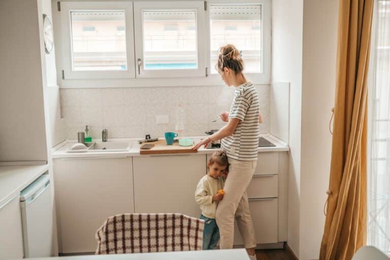 Mother standing at sink with toddler hugging her legs