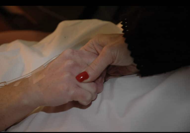 Two women holding hands over a hospital bed, color photo