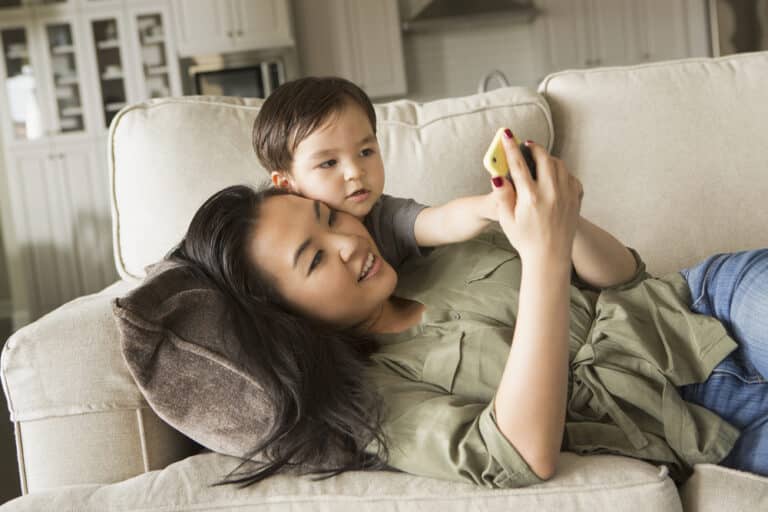 Woman lying on couch looking at smartphone with child over her shoulder