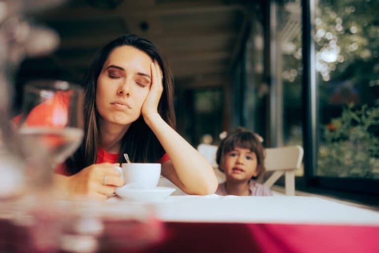 Tired mother with coffee cup on table, child sitting next to her