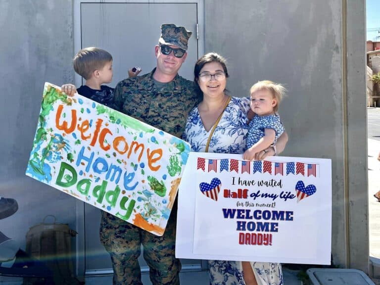 Mother and two children holding "welcome home" signs next to soldier daddy, color photo