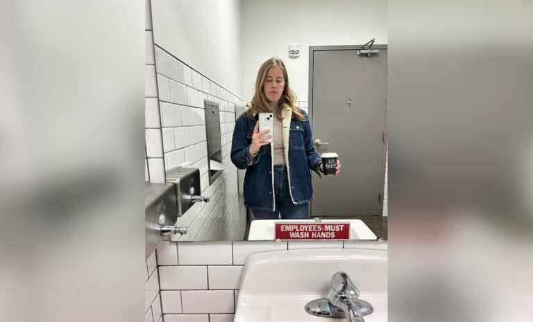 Woman taking a selfie in a bathroom mirror holding a coffee cup