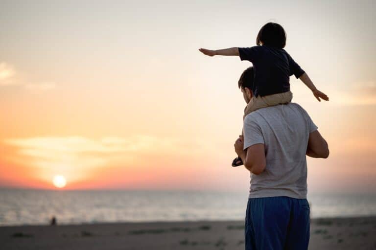 Son on dad's shoulders looking at sunset over water