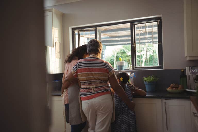 Mother, daughter, and grandmother stand at sink, view from behind