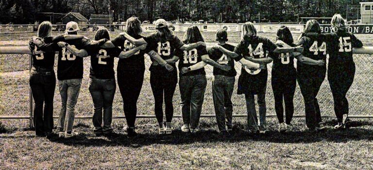 Moms with arms around each other watching baseball